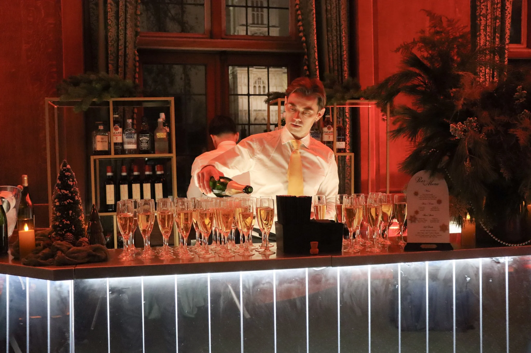 A bartender pouring a drink into champagne glasses at a festive bar setup with Christmas decorations and bottles of liquor in the background.