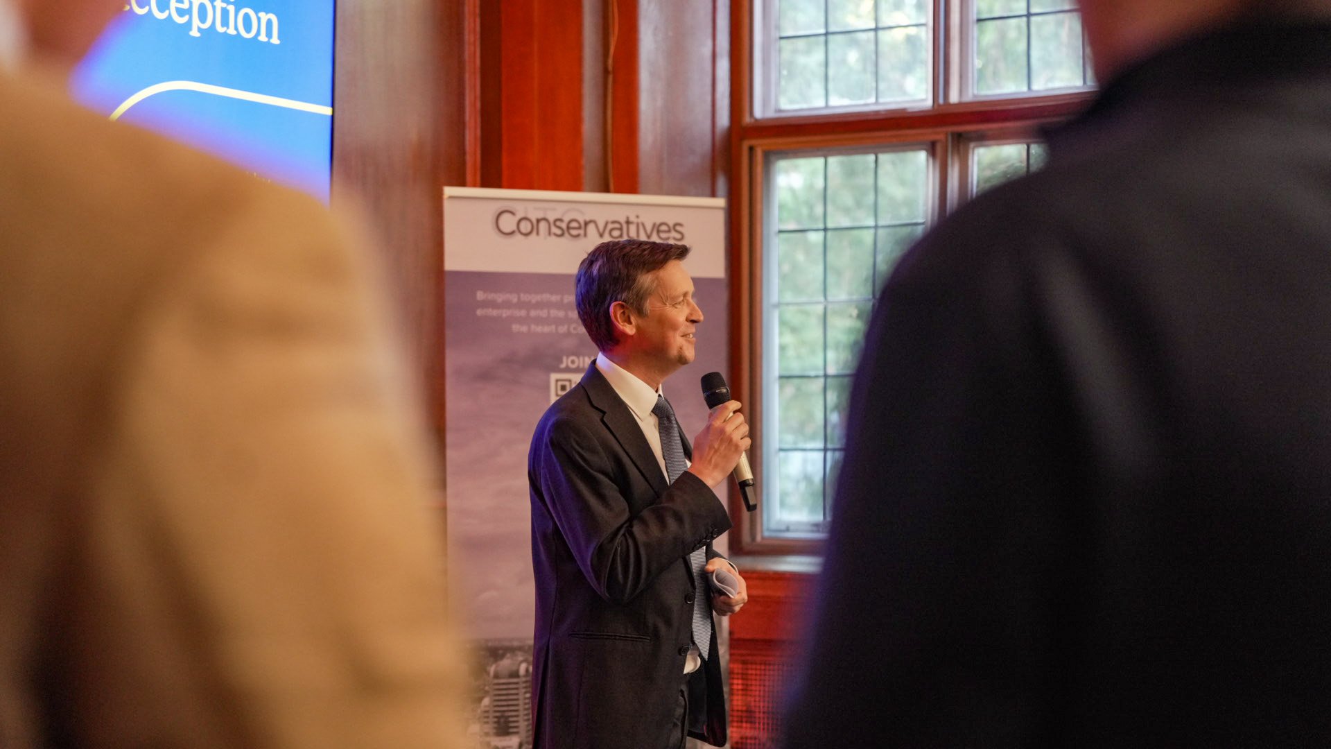 A man in a dark suit and tie speaking into a microphone at a political event, with a banner behind him that reads 'Conservatives'. The background features wooden walls and large windows.
