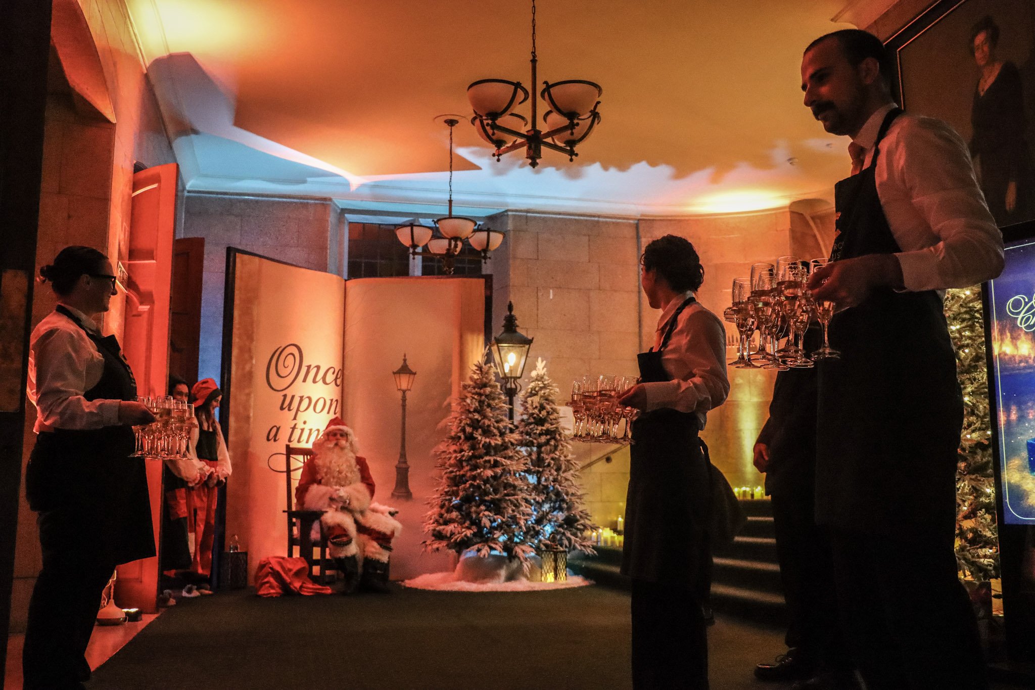 Indoor holiday scene with Santa Claus sitting in front of a small decorated Christmas tree, three women serving drinks, a Santa figurine, and festive Christmas decorations. Warm lighting and a sign that reads 'Once upon a time' are visible.