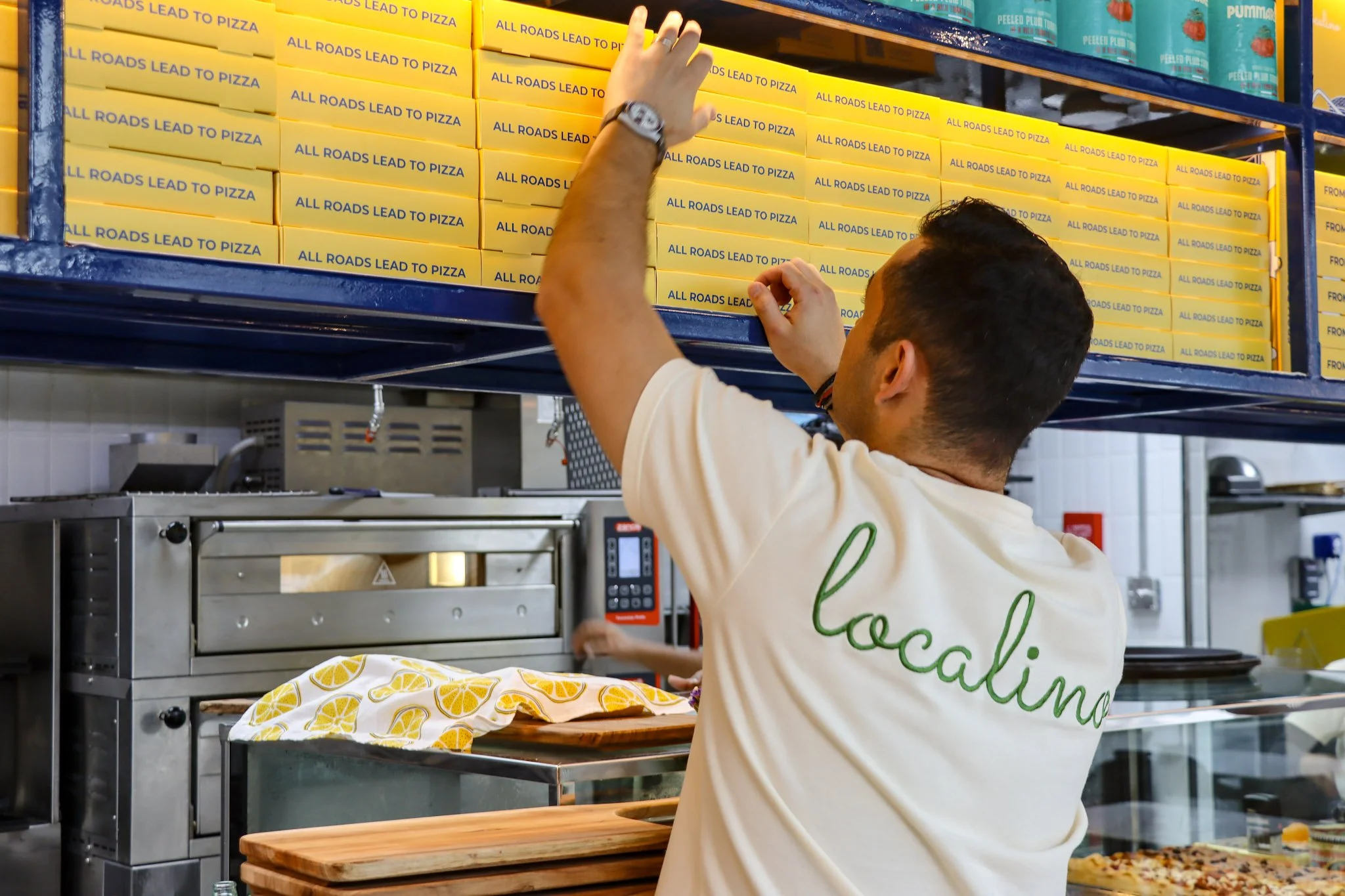 A person reaching for a yellow pizza box in a store aisle filled with pizza boxes labeled "All Roads Lead to Pizza."