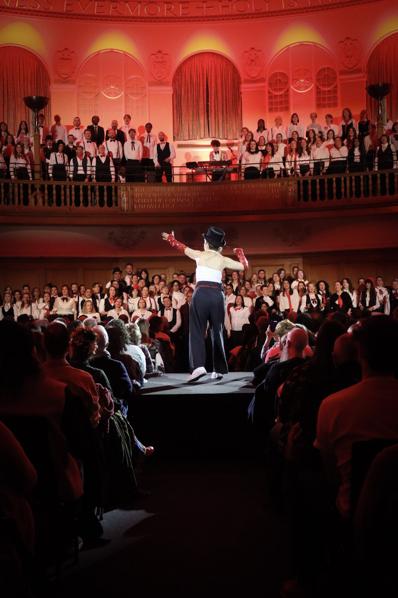 Choir performing on stage in a historic concert hall. The conductor is standing in front, facing the choir. The choir is dressed in coordinated outfits with red accents and is arranged on tiered risers. The hall features ornate decorations and warm lighting.