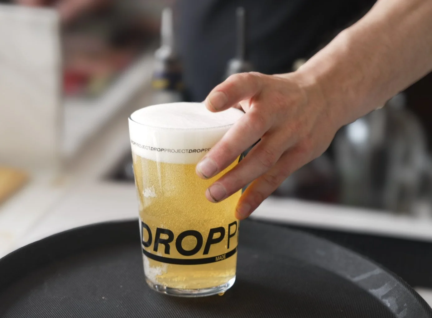 A person's hand placing a pint glass of beer on a black tray.