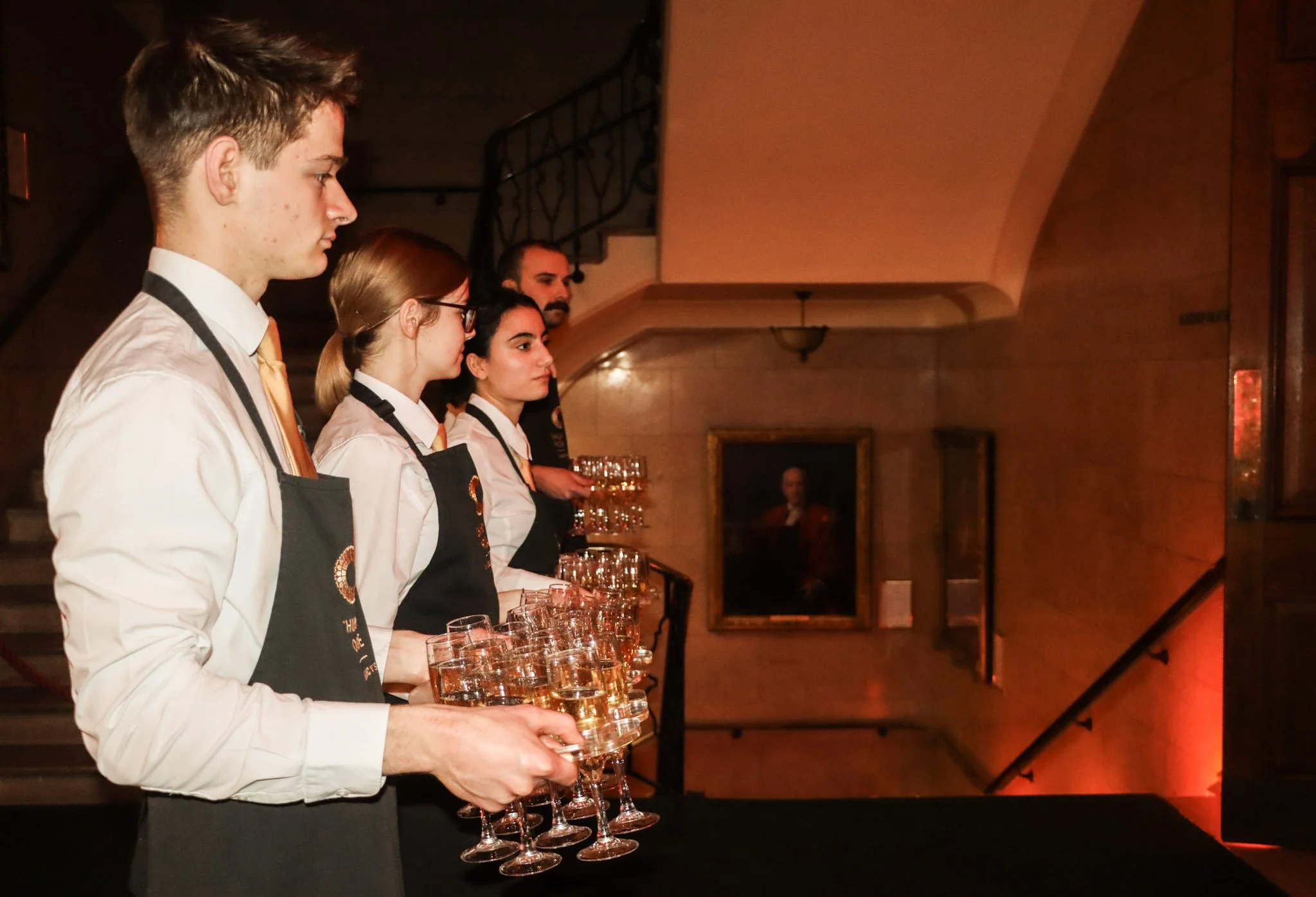 Four people in white shirts and black aprons lined up in a row, each holding multiple glasses of water, during a formal event or ceremony in a dimly lit room with wooden decor and framed pictures on the walls.