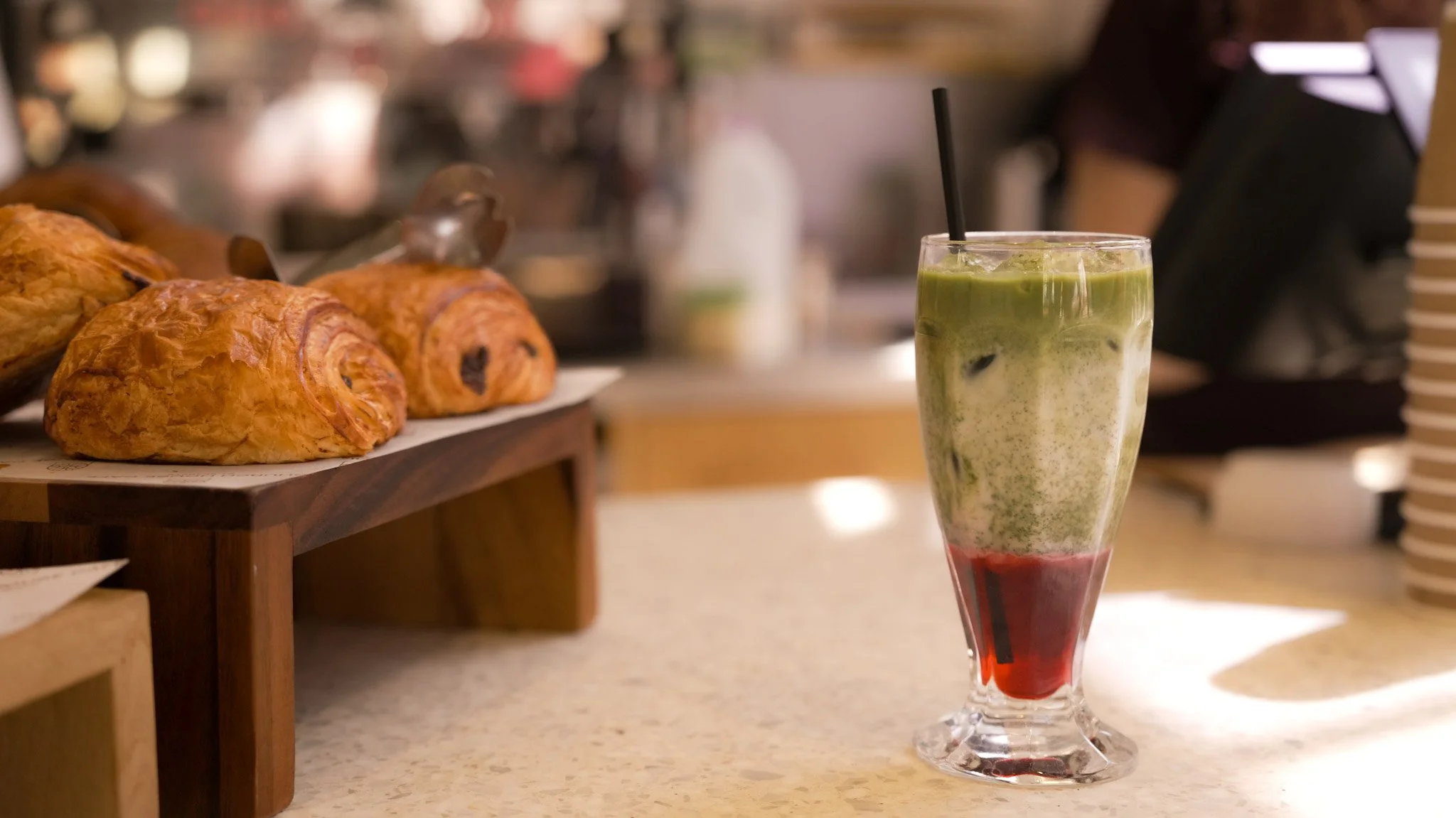 A layered milkshake with green, white, and red layers in a tall glass with a black straw, placed on a counter in a cafe. In the background, croissants are displayed on a wooden tray.