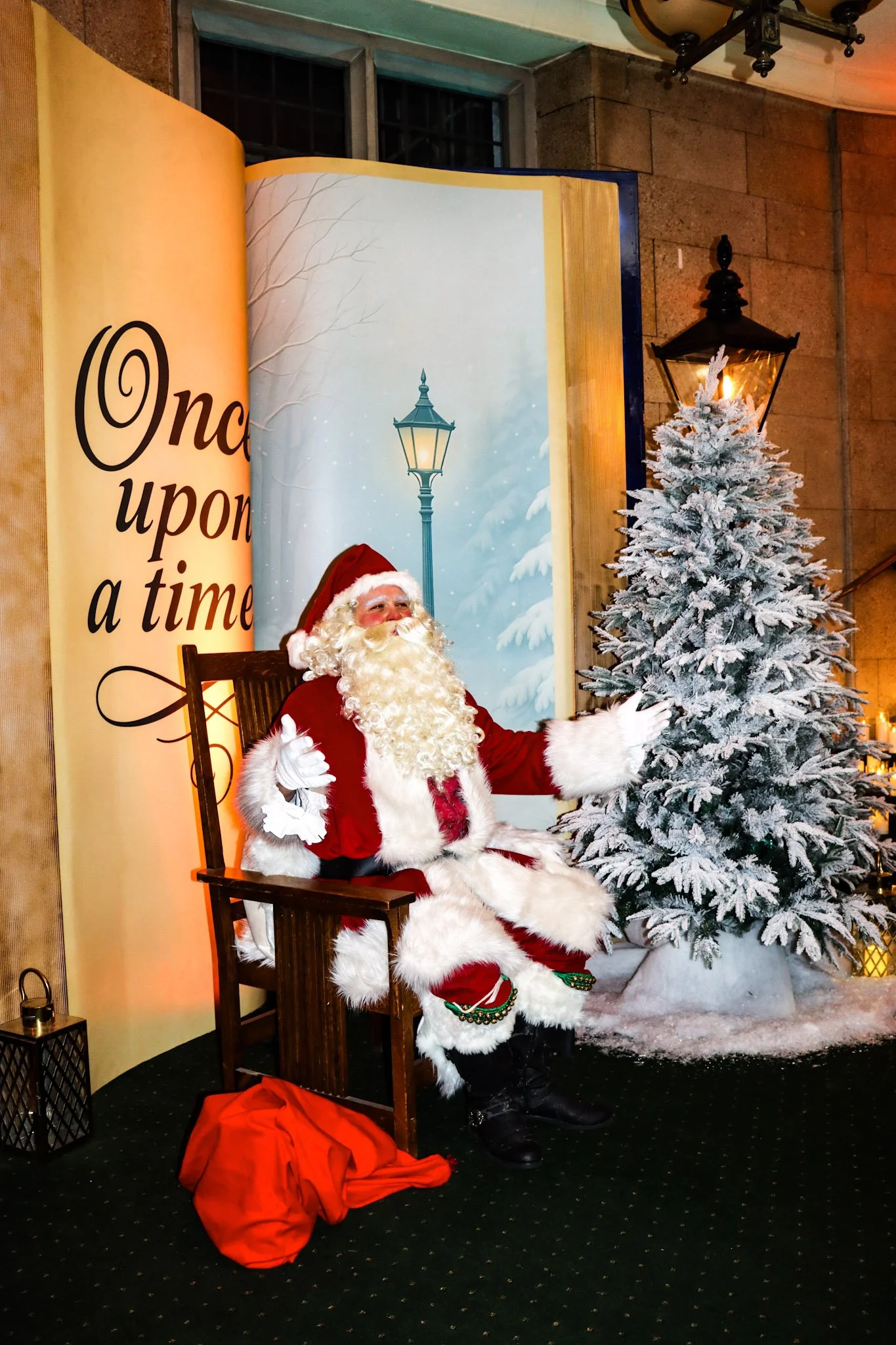 Santa Claus wearing a red and white suit, sitting on a wooden chair next to a snow-covered Christmas tree with a lit candle on top. The background features a winter scene with a streetlamp and snow, with a banner on the left that reads 'Once upon a t