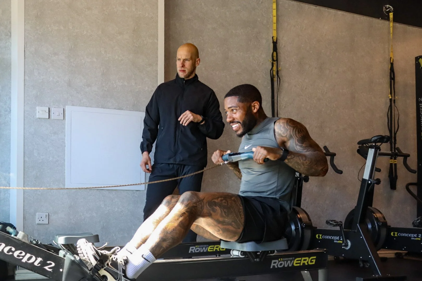 A man with tattoos on his legs and arms is seated on a rowing machine, smiling and pulling the handles while a trainer watches. The trainer is standing nearby, observing him. The room has fitness equipment and resistance bands mounted on the wall.