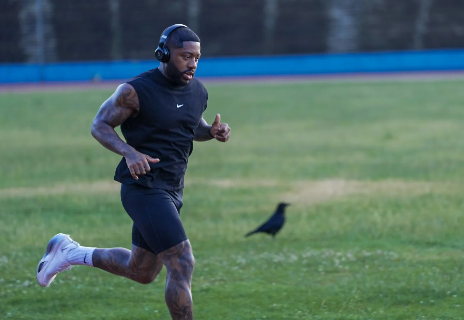 A man running outdoors on a grassy field wearing a black sleeveless shirt, black shorts, white sneakers, and headphones, with a black bird in the background.