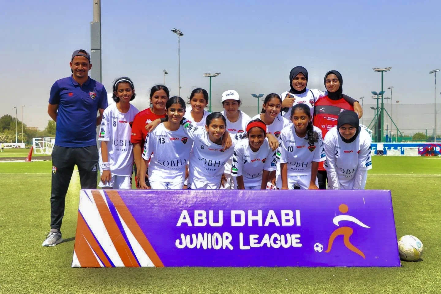 Group of young female soccer players and coaches posing on a soccer field with a purple sign that reads 'Abu Dhabi Junior League' in front of them.