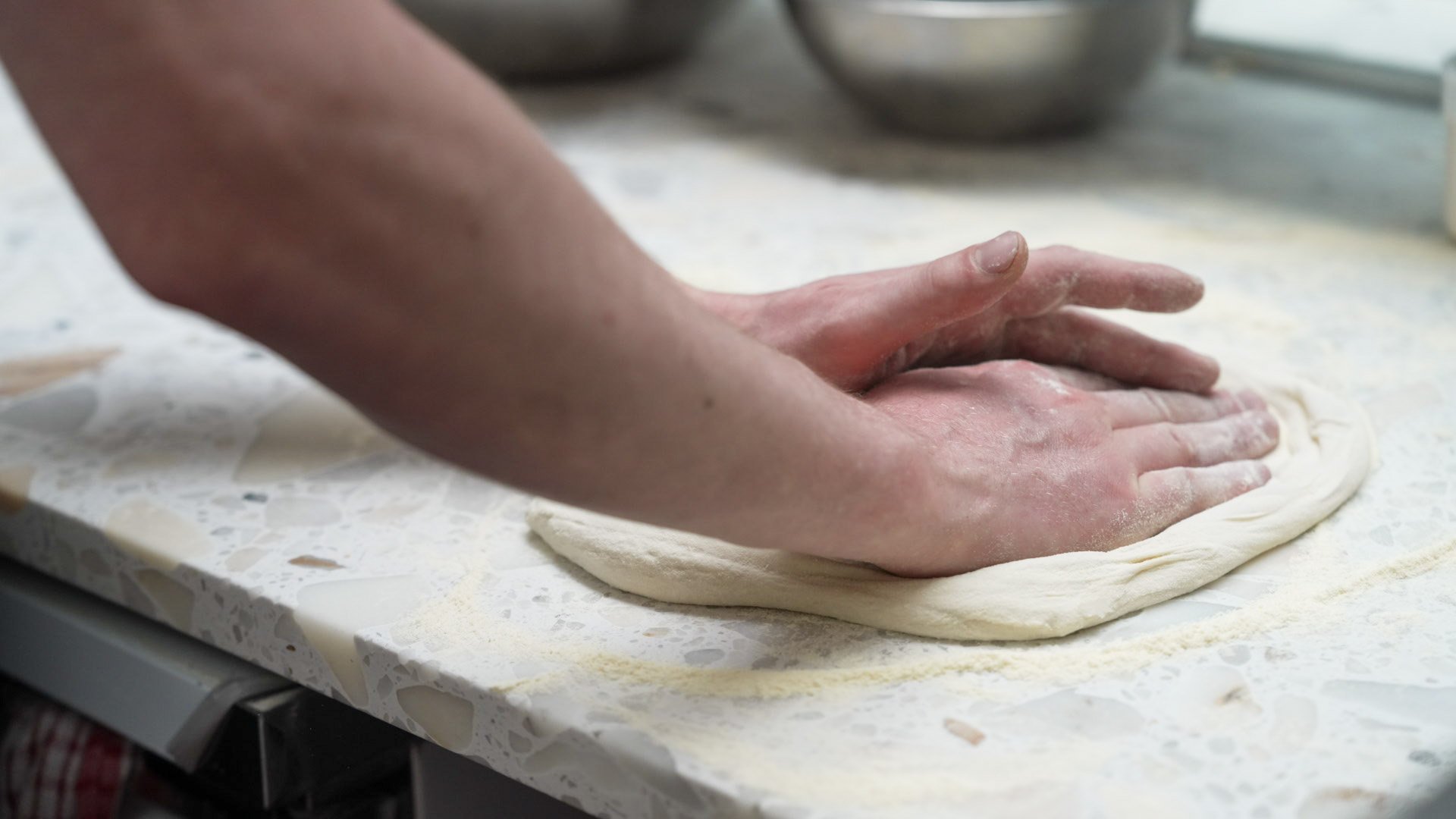 Person kneading pizza dough on a marble surface in a kitchen.