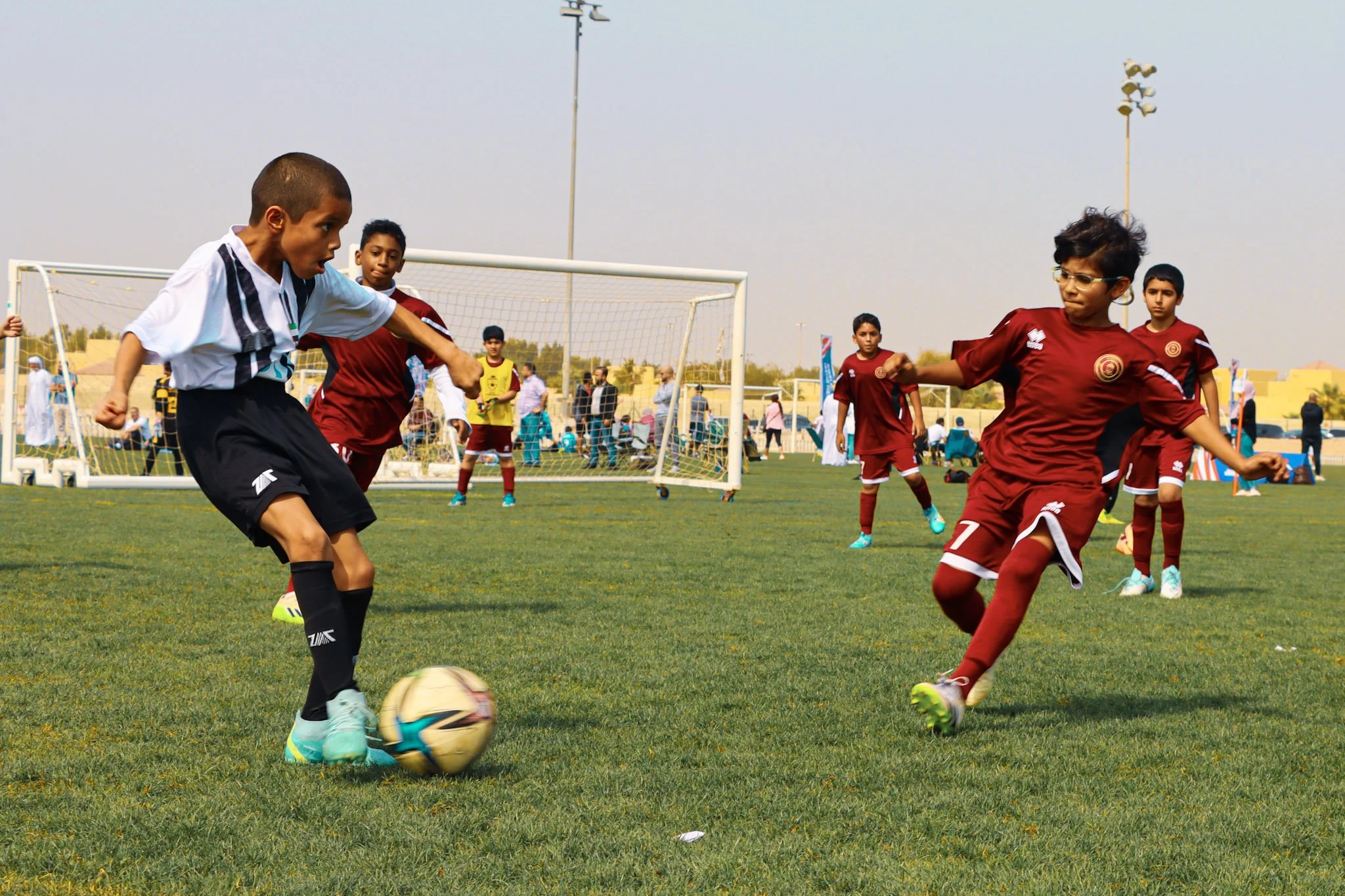 Children playing soccer on a field with goalposts and spectators in the background.