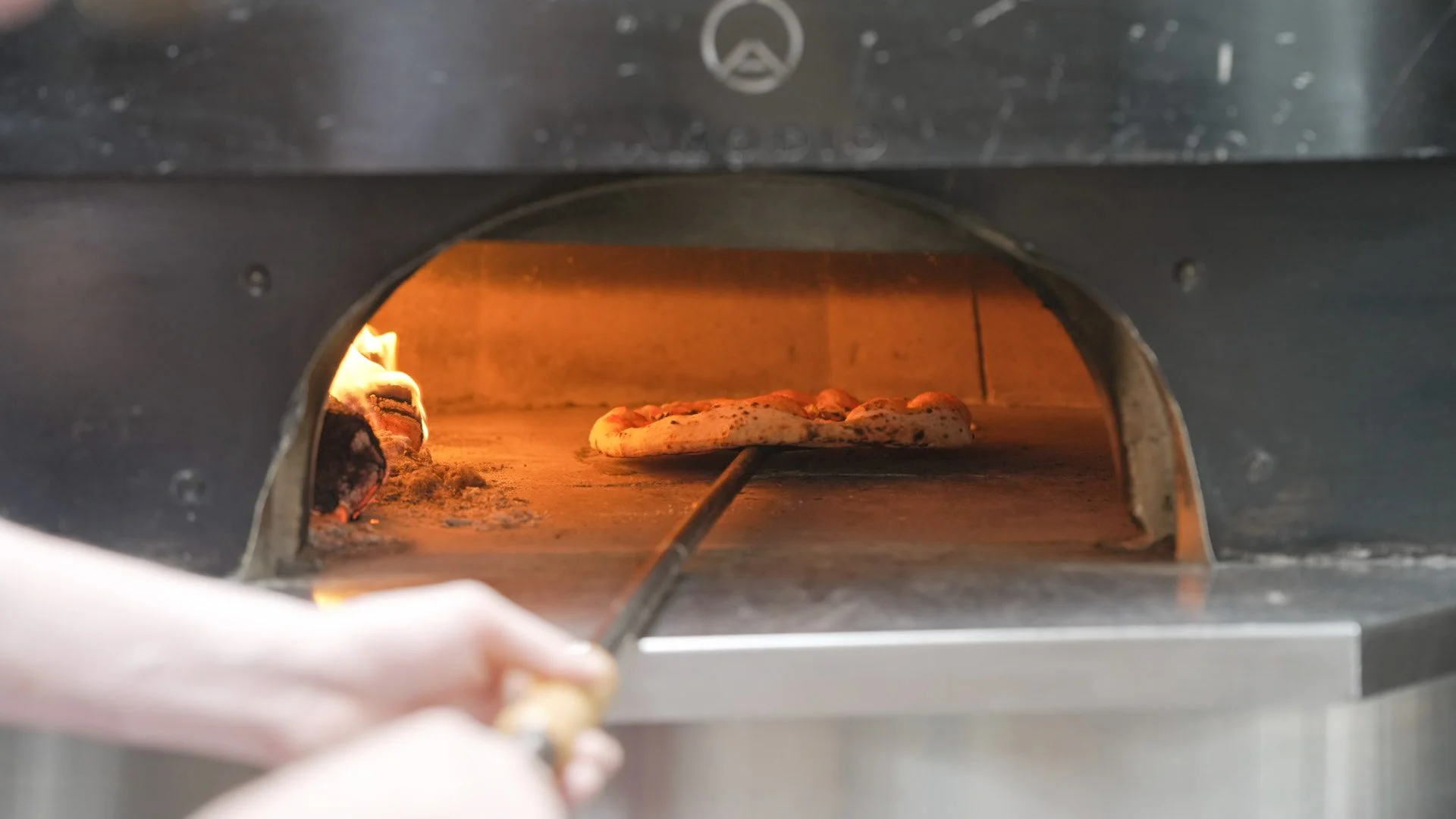 A pizza baking in a wood-fired oven, with a person using a pizza peel to place or remove it.