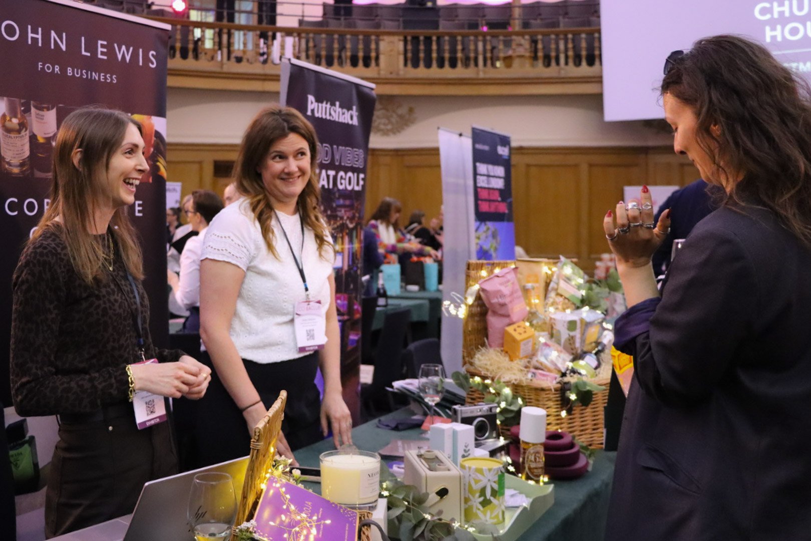 Three women at a booth conversing at an indoor event. The woman on the left has long brown hair and is wearing a dark animal print top, smiling. The woman in the middle has wavy brown hair, wearing a white top, smiling. The woman on the right has dar