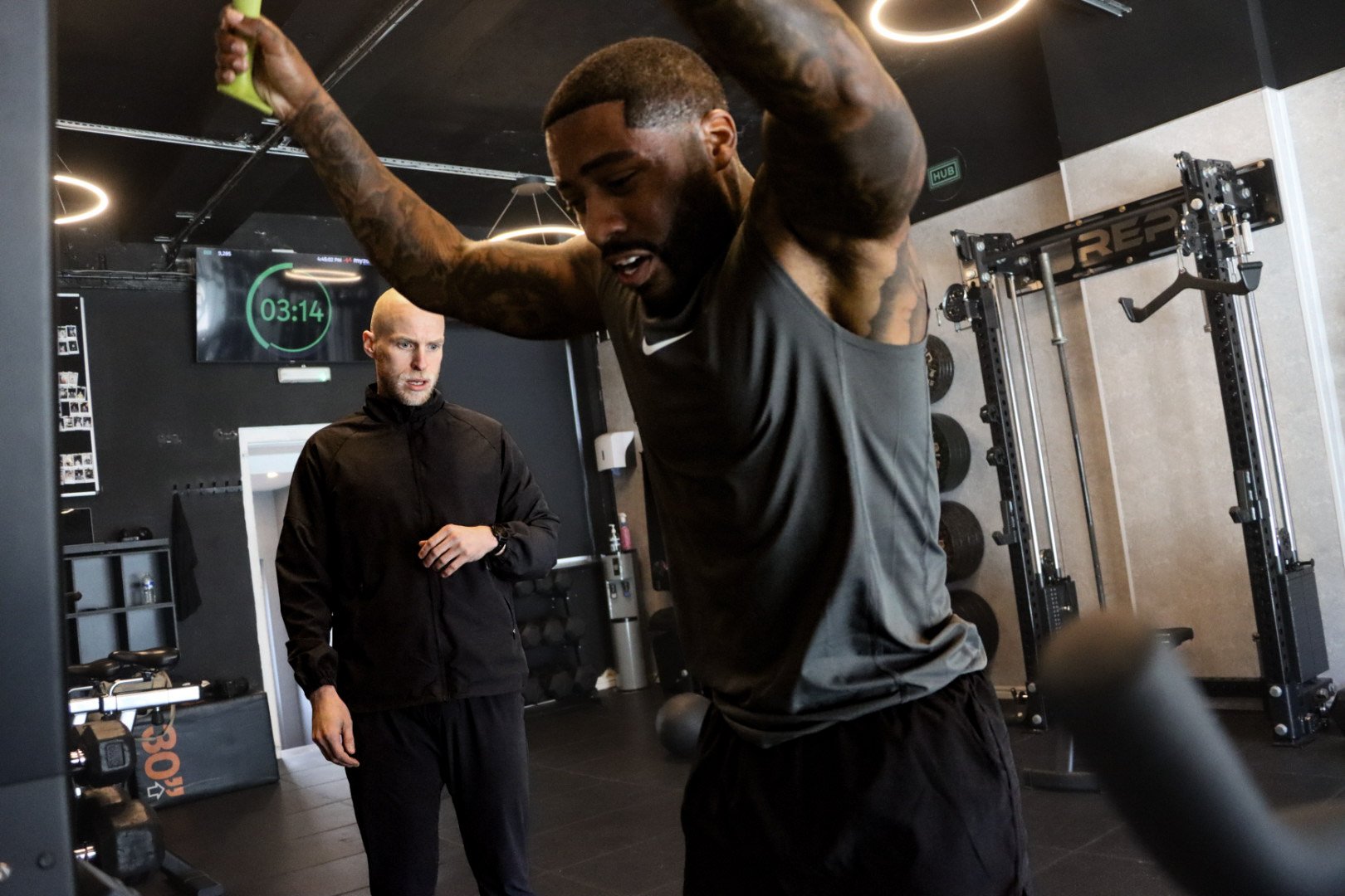 A man is doing a workout in a gym while a trainer observes him. The man is lifting a barbell with weights. The trainer is standing nearby, watching. There is a digital timer on the wall showing 3 minutes and 14 seconds.