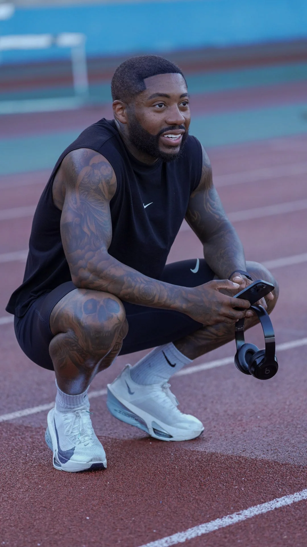 A man crouching on a running track holding a phone in his hands, with headphones hanging from his neck, wearing a black sleeveless T-shirt, black shorts, white athletic shoes, and white socks.