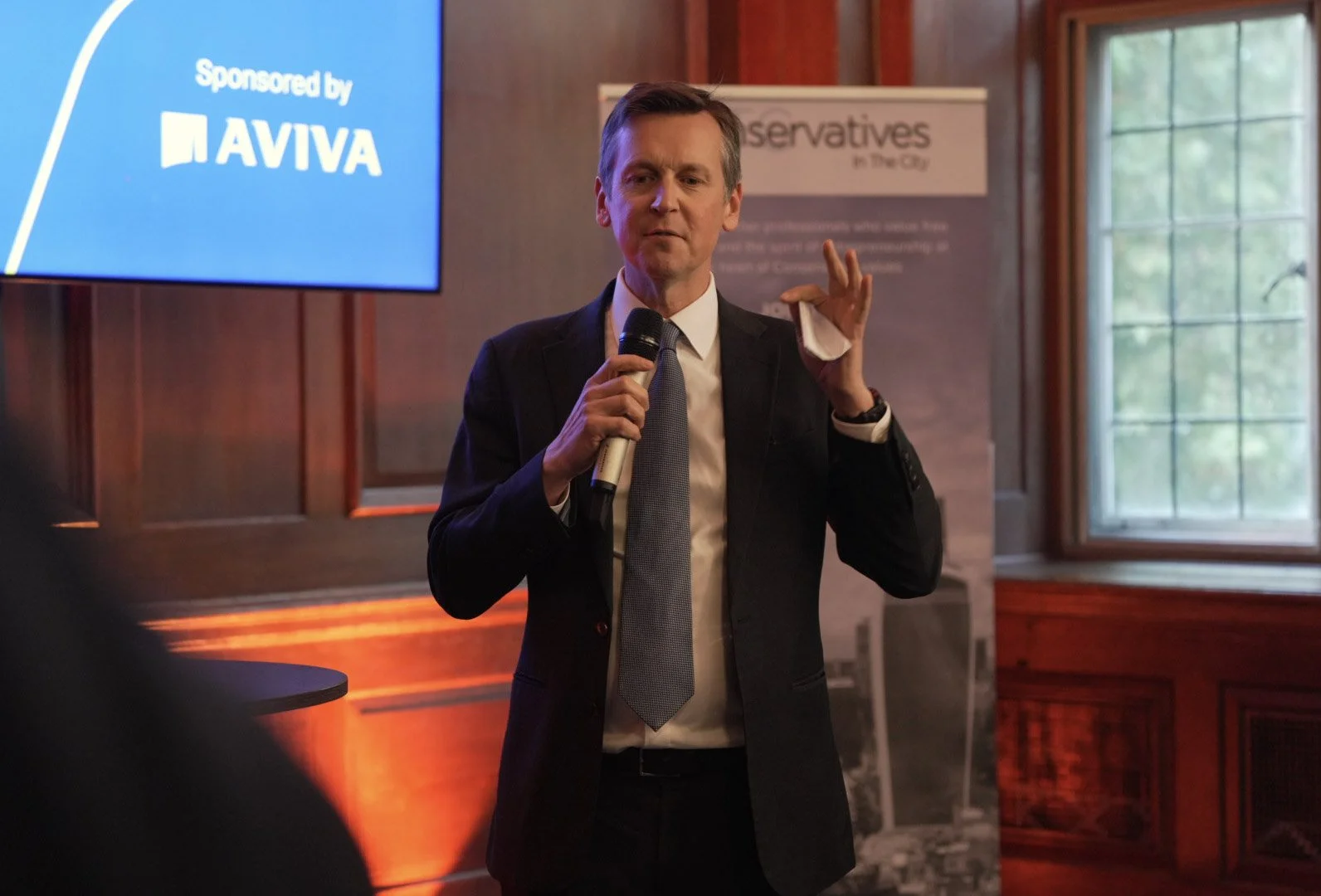 A man in a suit holding a microphone and speaking at a conference or event. A blue screen with the word 'Sponsored by AVIVA' is visible in the background.