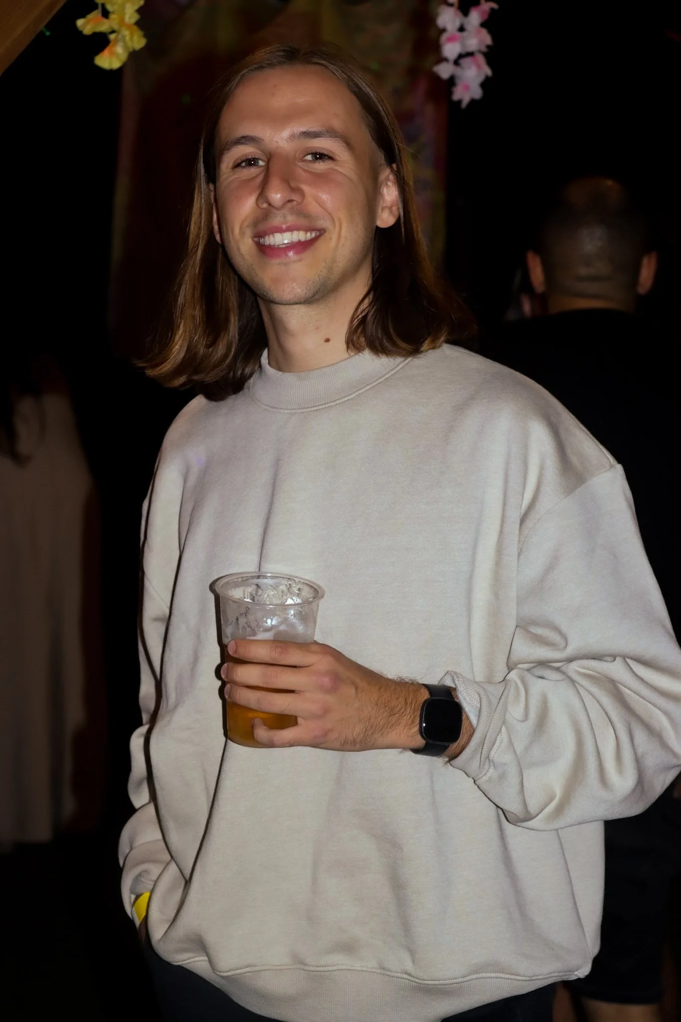 A young man with long brown hair smiling and holding a plastic cup of beer in an indoor setting with floral decorations in the background.