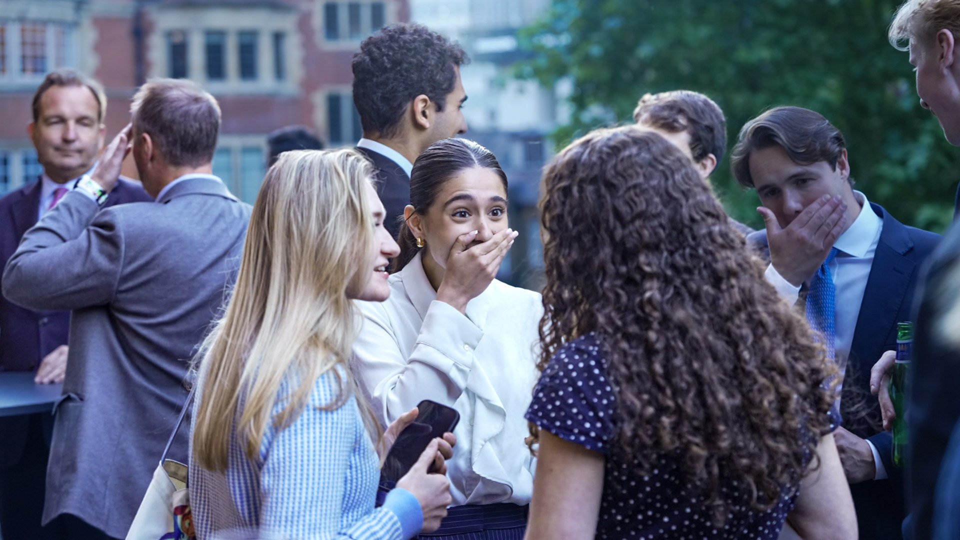 Group of people in formal attire at an outdoor event, some smiling and talking, with a woman covering her mouth in surprise or laughter, and others engaging in conversation.