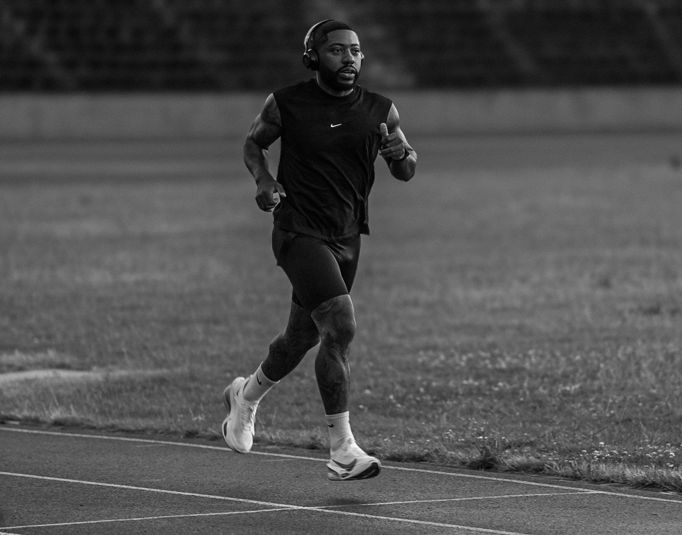 A man running on a track, wearing a sleeveless athletic shirt, shorts, and running shoes, with headphones on.