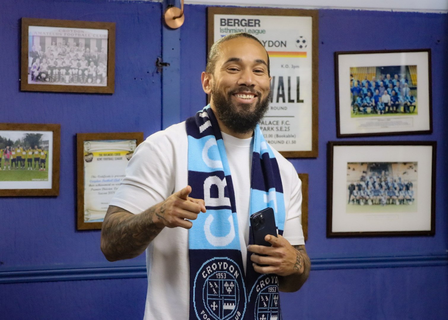 A man with a beard smiling and pointing at the camera, wearing a white shirt and a blue and white football scarf. He is standing in front of a blue wall decorated with framed football team photos and club memorabilia.