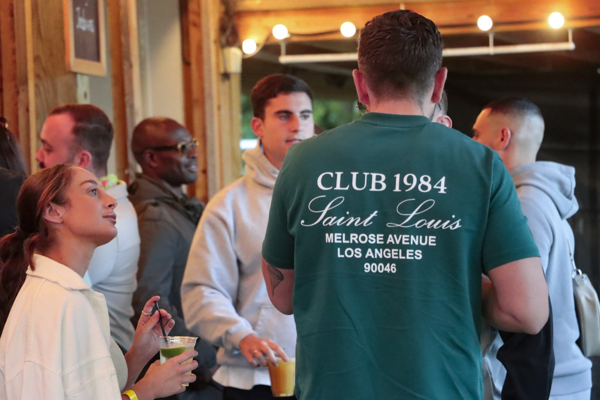 Group of people socializing at a bar or party, with a woman in a white shirt holding a drink and a straw in the foreground, and a man in a green shirt with 'CLUB 1984 Saint Louis' printed on the back standing with his back to the camera.