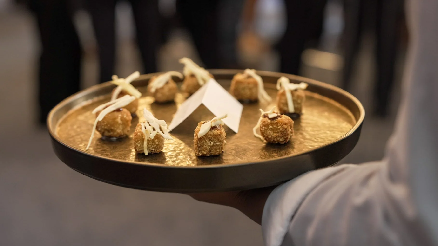 Tray of fried appetizer bites garnished with shredded cheese, held by a person in a white shirt, with a blurred background.