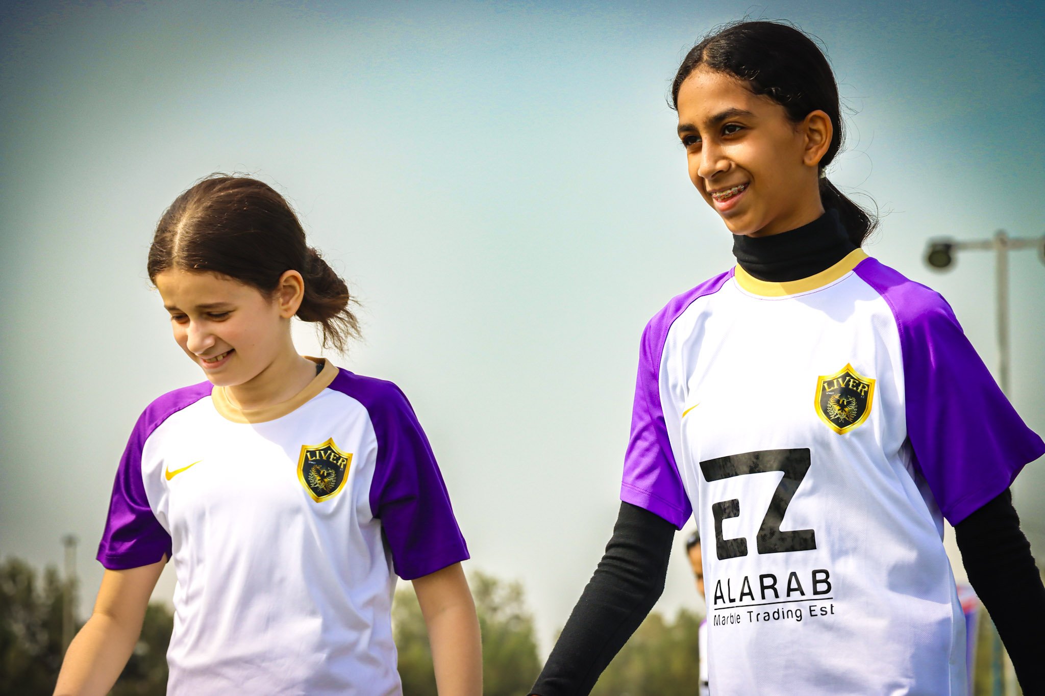Two young female soccer players wearing white and purple jerseys walk hand-in-hand on the field during daytime, smiling and enjoying the game.
