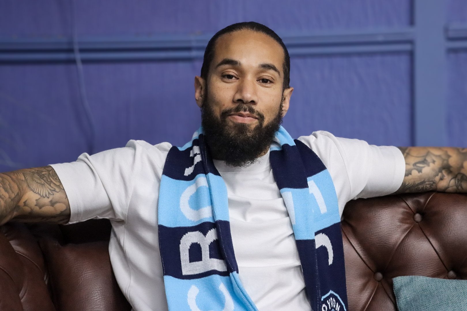 A man with a beard and tattoos on his arms sitting on a brown leather couch, wearing a white t-shirt and a blue and white scarf, with a blue background behind him.