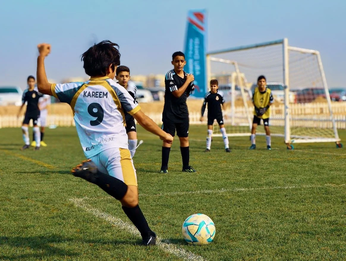 Young soccer player with jersey number 9 named Kareem preparing to kick a soccer ball during a game on a grass field, with teammates and a goalkeeper in the background.