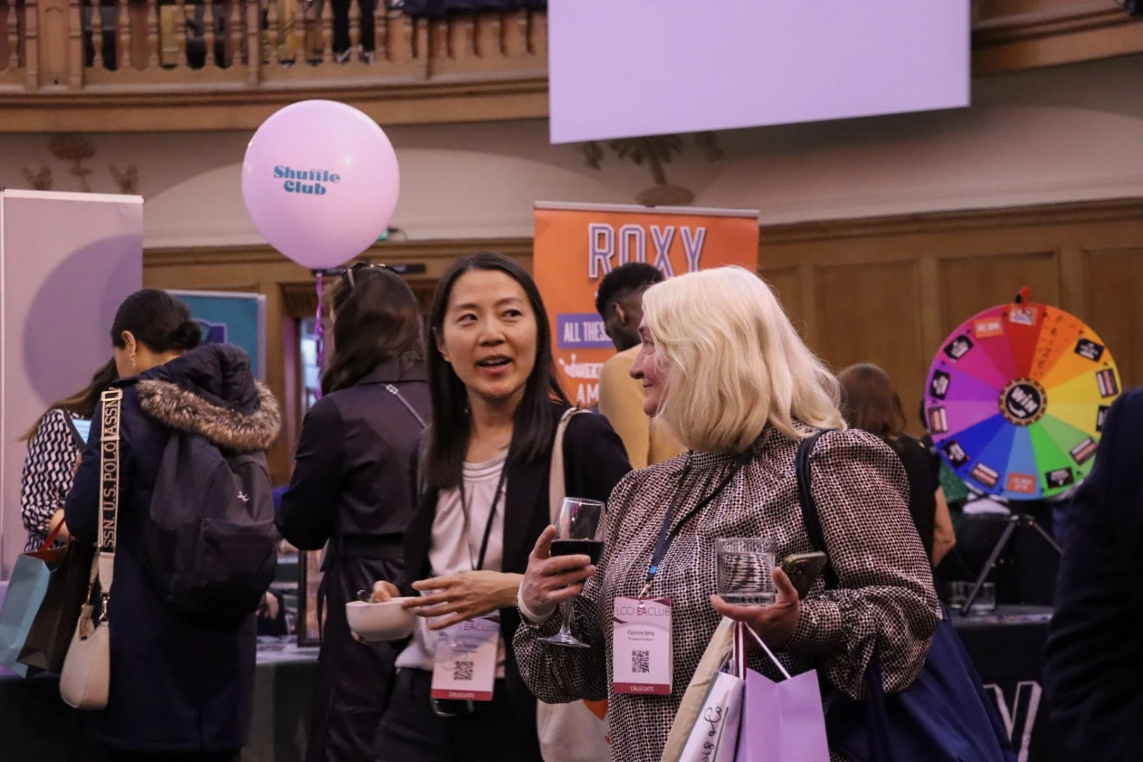 People socializing at an event, with one woman holding a glass of red wine and another holding a glass of water, surrounded by event booths and colorful signage.