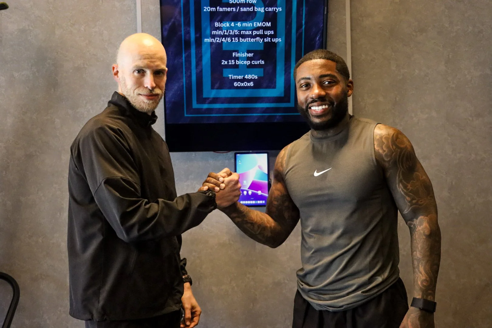 Two men shaking hands in a gym or fitness center, with workout and training information displayed on a screen behind them.