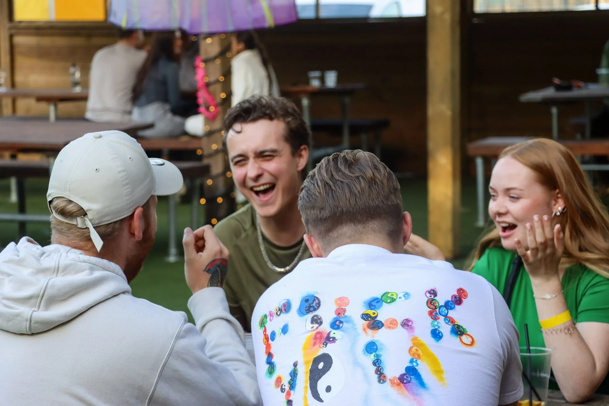 Group of young people laughing and socializing outdoors at a party, with a picnic table, wooden walls, and string lights in the background.