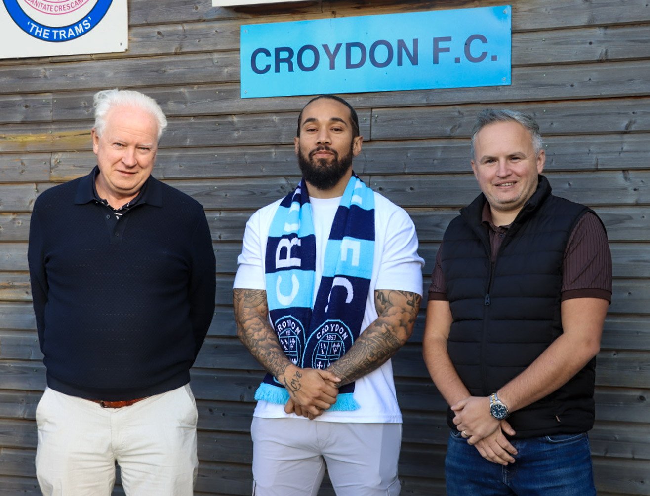 Three men standing in front of a wooden wall with a blue sign that reads 'CROYDON F.C.'. The man in the middle is wearing a scarf with the Croydon F.C. logo, has tattoos on his arms and a beard. The man on the left has white hair, is wearing a black 