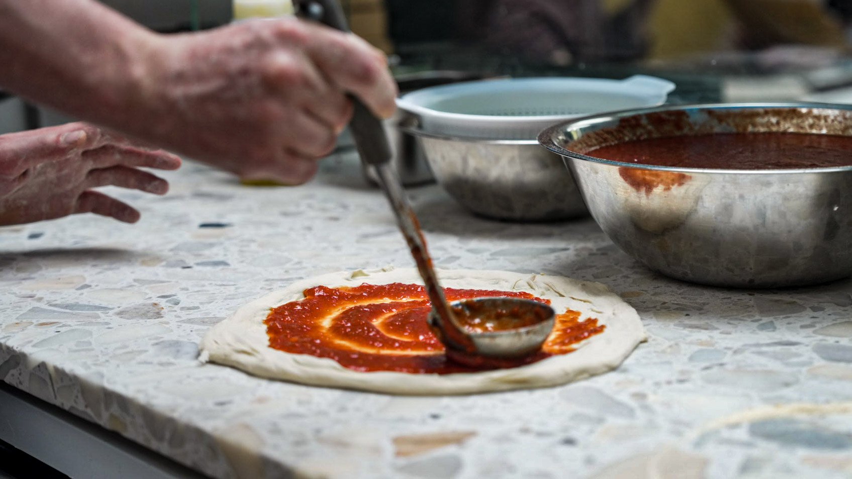 Hands spreading tomato sauce on pizza dough with a ladle, with bowls of sauce and other ingredients on a kitchen countertop.