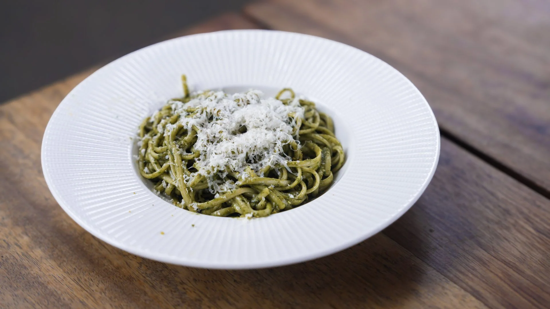 Plate of green pesto pasta topped with grated white cheese on a wooden table.
