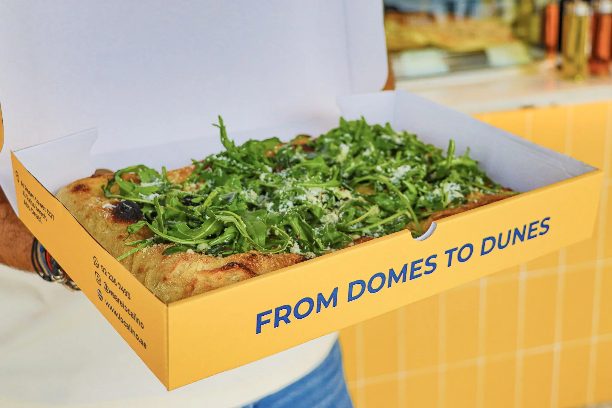Pizza with arugula and cheese in a yellow takeout box.