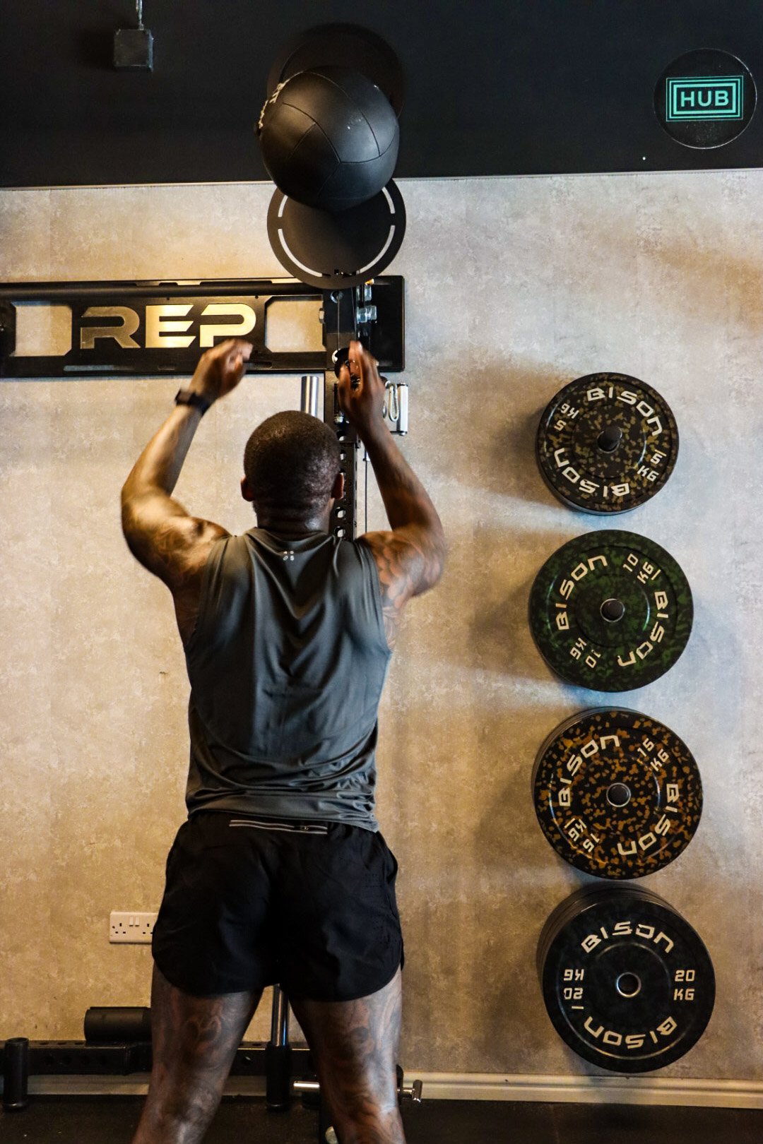 A man with muscular arms and tattoos is performing a workout at a gym, using a pull-up station with a black medicine ball hanging on top. There are weight plates mounted on the wall behind him and an illuminated sign reading 'REPS' above the equipmen