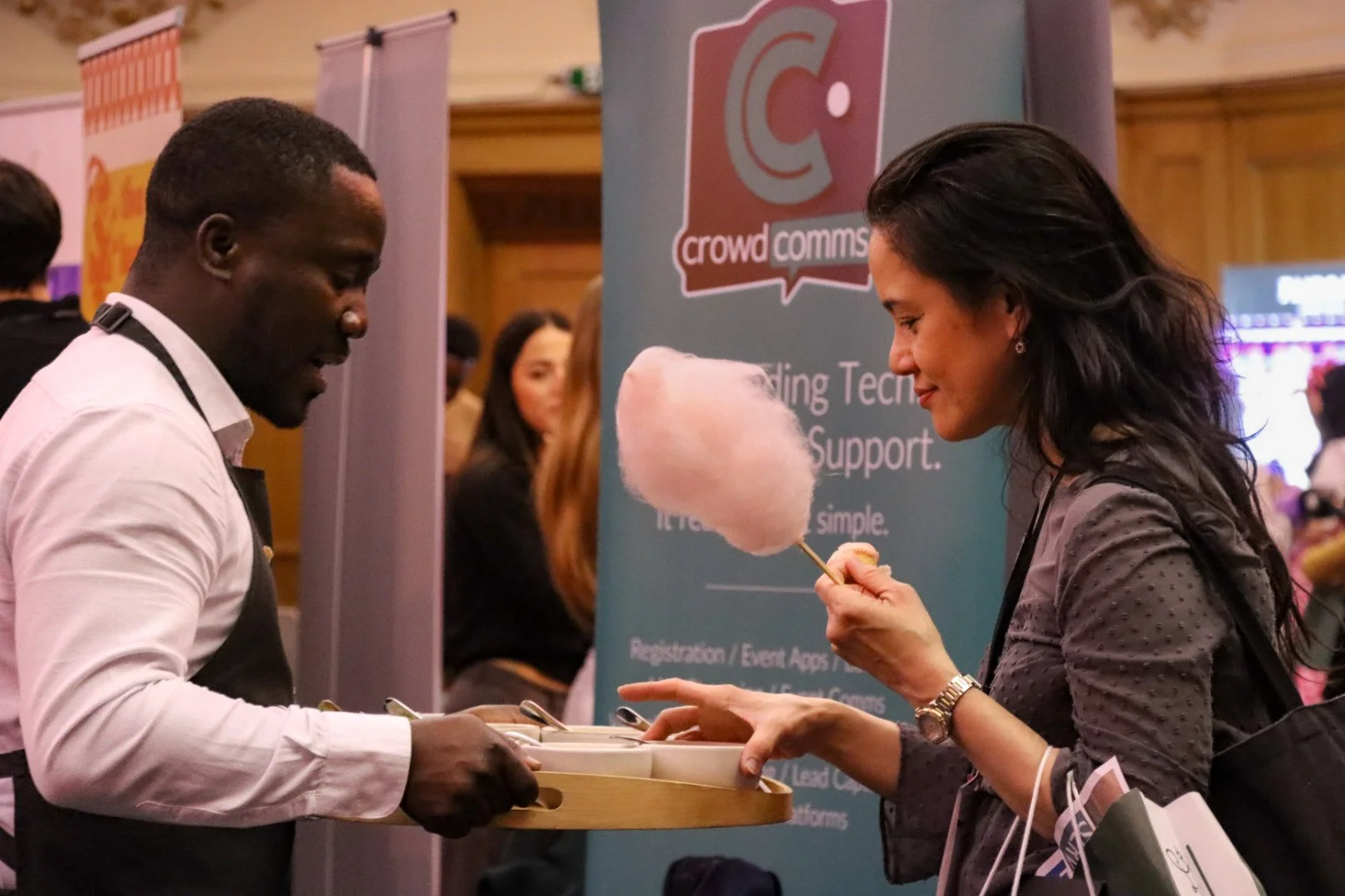 A woman with dark curly hair is holding pink cotton candy on a stick, smiling, and reaching for a tray held by a man. The man, with short black hair, is wearing a white shirt and a black apron. They are at a crowded event or conference, with a blue b
