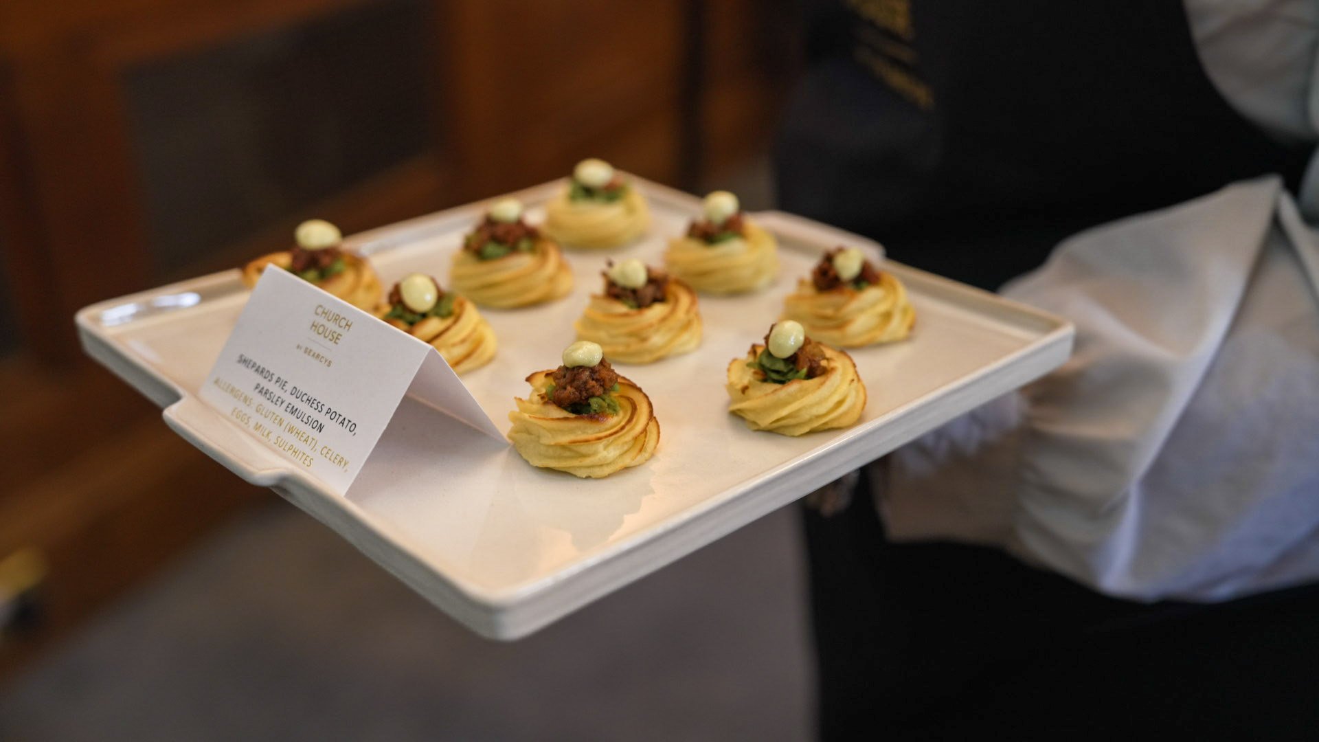 Tray of bite-sized appetizers topped with dollops of white sauce, ground meat, and green herbs, labeled as 'Shemard's Pie Duchess Potato, Parsley Emulsion' at a gathering or event.
