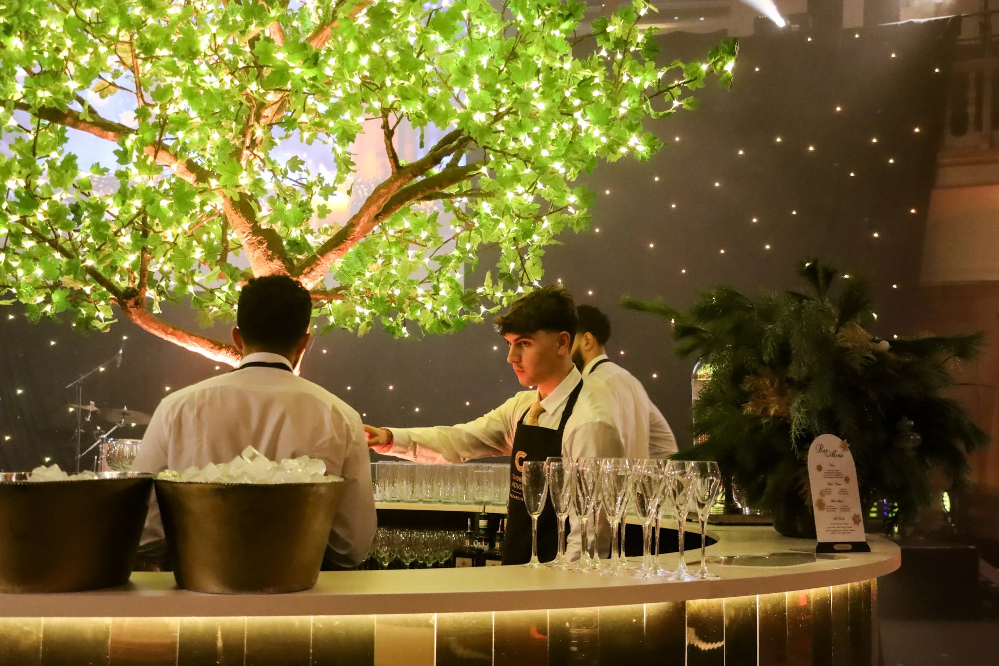 Bar with bartenders preparing drinks, illuminated green leaves on a tree, and a dark ceiling with small star-like lights at a fancy event or restaurant.