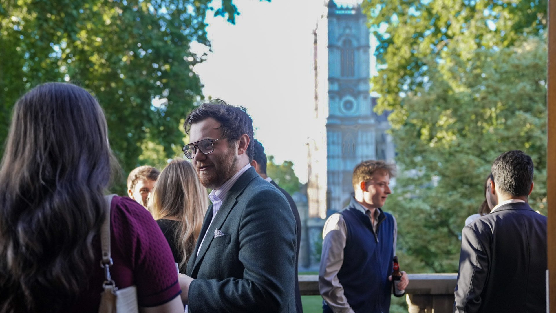 Group of young adults socializing outdoors in a park during daytime, with trees and a historic building with a clock tower in the background.