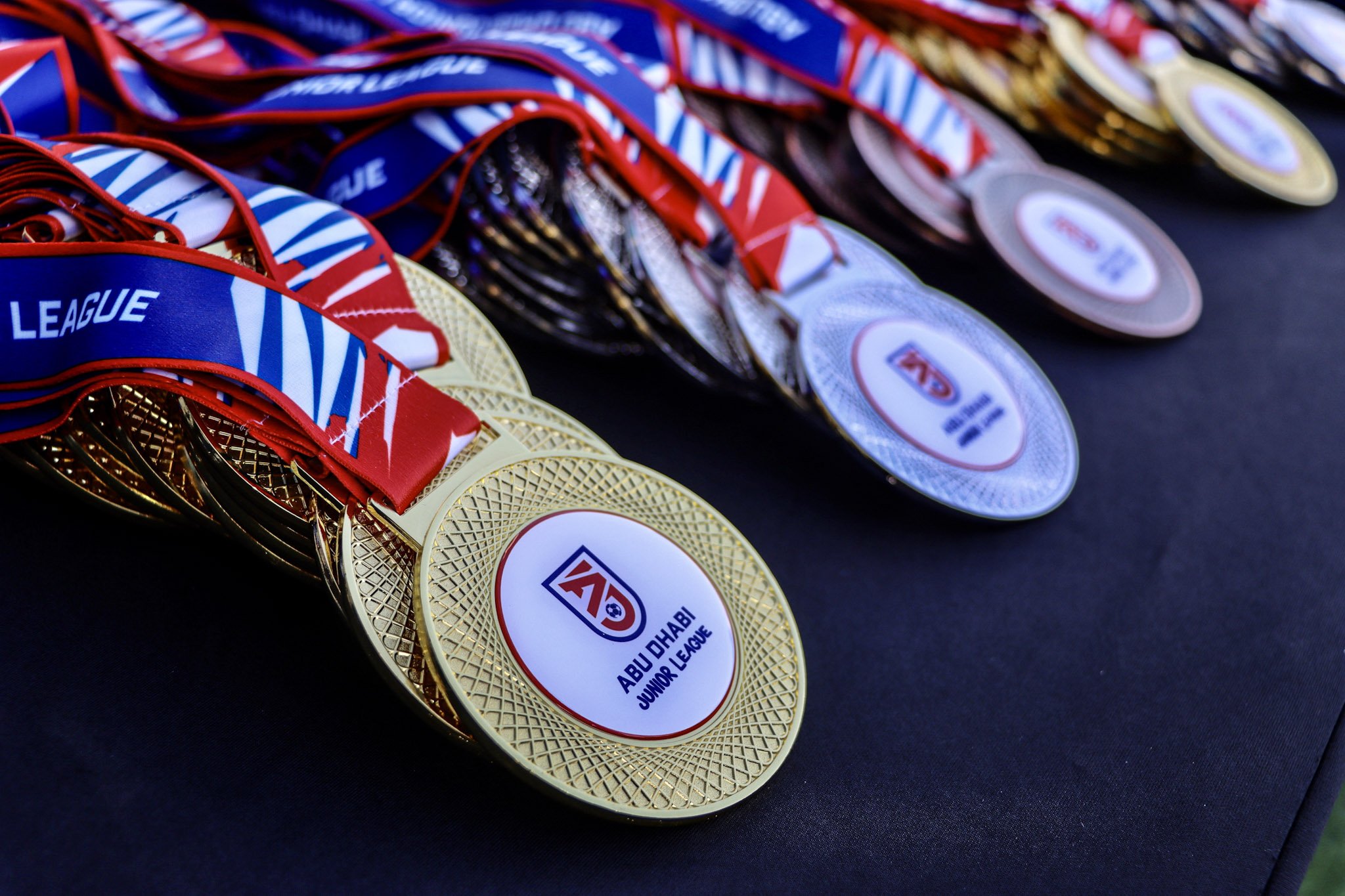 Close-up of medals for Abu Dhabi Junior League with red, white, and gold ribbons, displayed on a black surface.