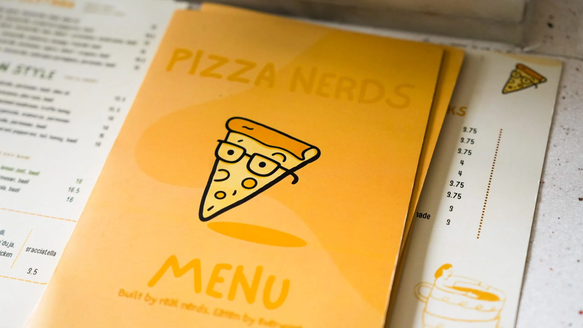 A yellow menu folder with the words 'Pizza Nerds' and a cartoon pizza slice with glasses on it. The menu is on top of a paper menu with prices.