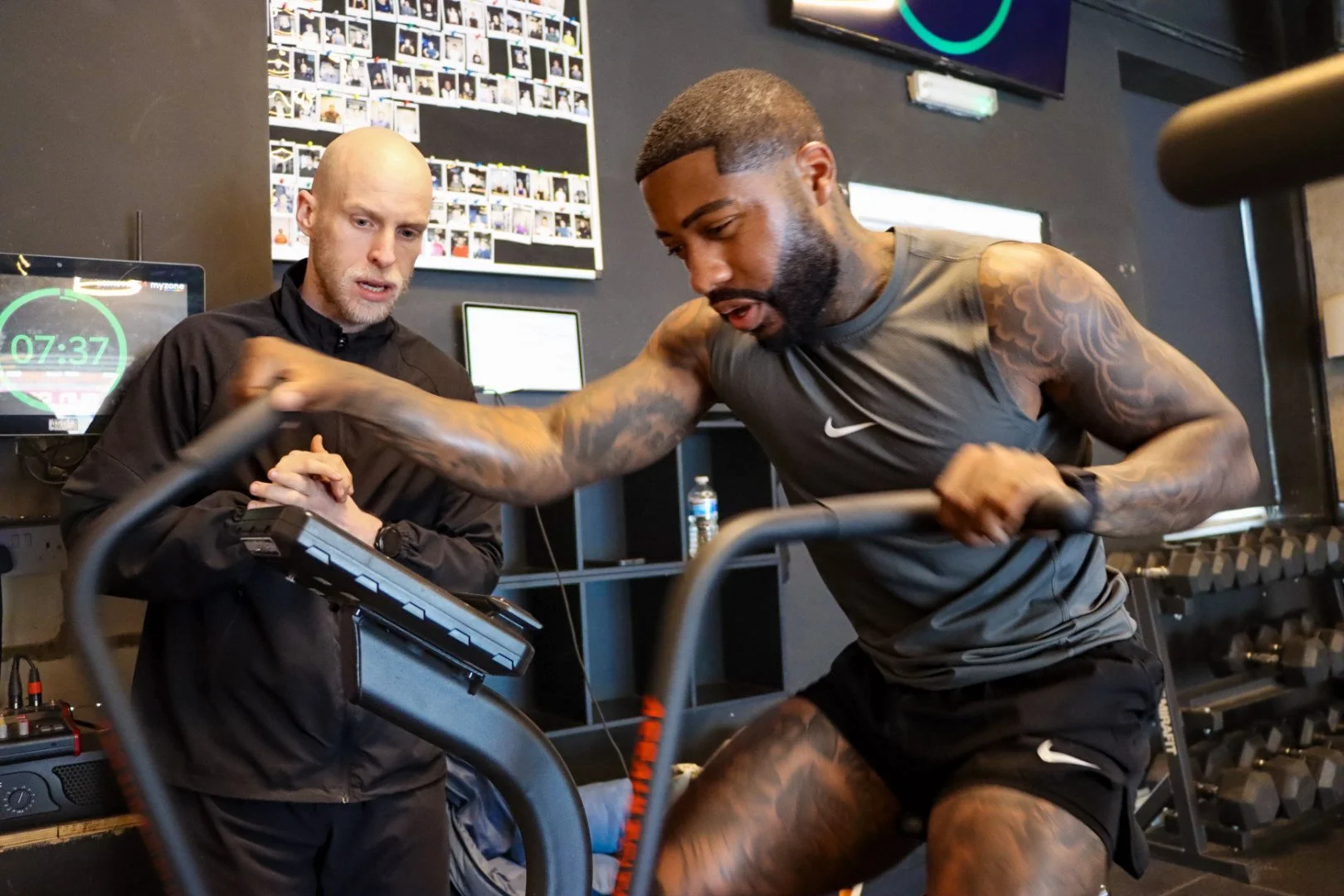 A muscular man with tattoos running on a treadmill while a trainer assists him in a gym.