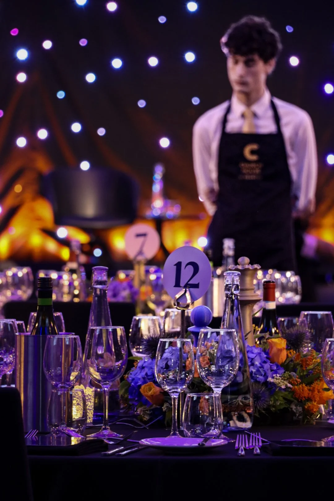 A banquet table setup with wine glasses, flowers, and table cards numbered 7 and 12, with a young man dressed in a shirt, tie, and apron standing behind it, in a dimly lit setting with colorful lights.