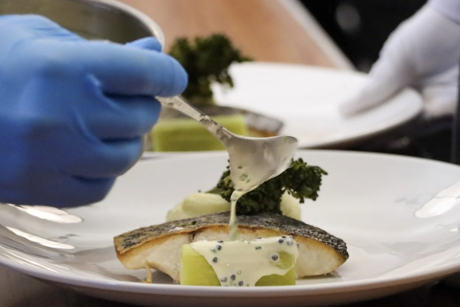 A chef is pouring a creamy sauce over a piece of cooked fish on a plate, garnished with vegetables like broccoli and a celery stalk, with another plate and chef in the background.