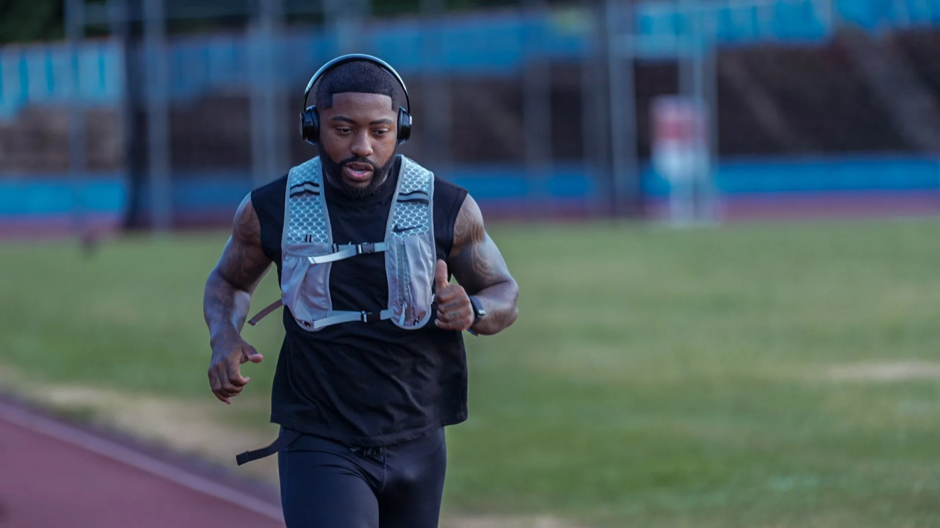A man running on an outdoor track, wearing headphones and a hydration vest.