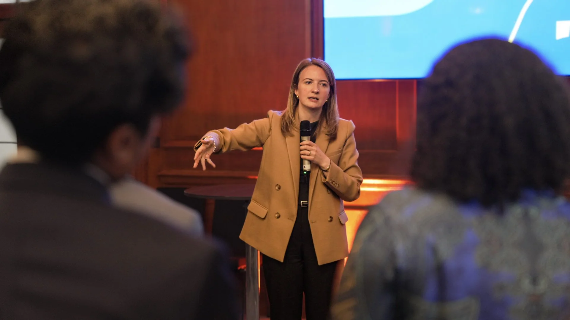 A woman in a tan blazer speaking into a microphone while giving a presentation in front of a large screen, with blurred audience members in the foreground.