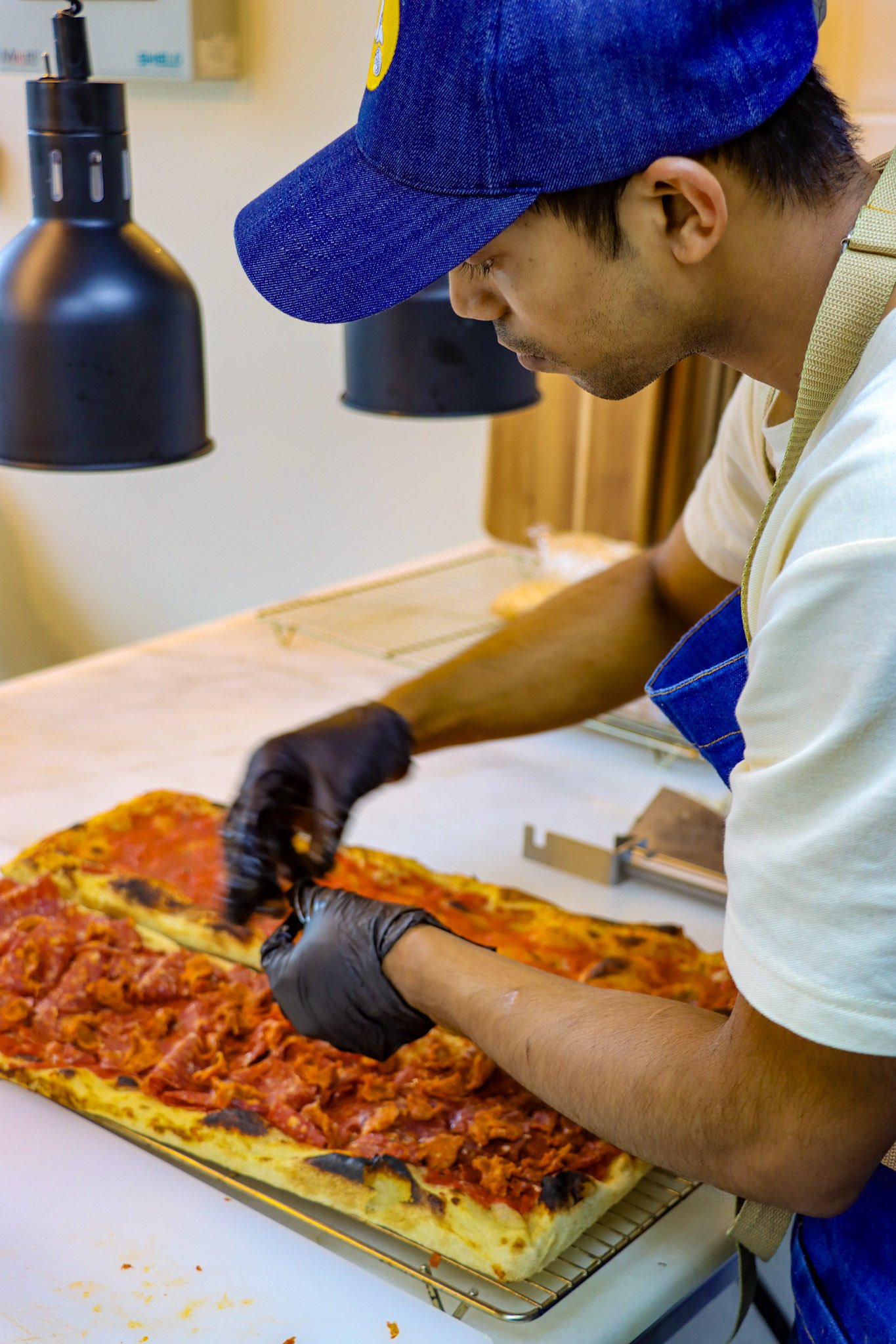 A man wearing a blue cap, a white shirt, an apron, and black gloves is slicing a freshly baked pizza with tomato sauce and toppings on a wire rack.