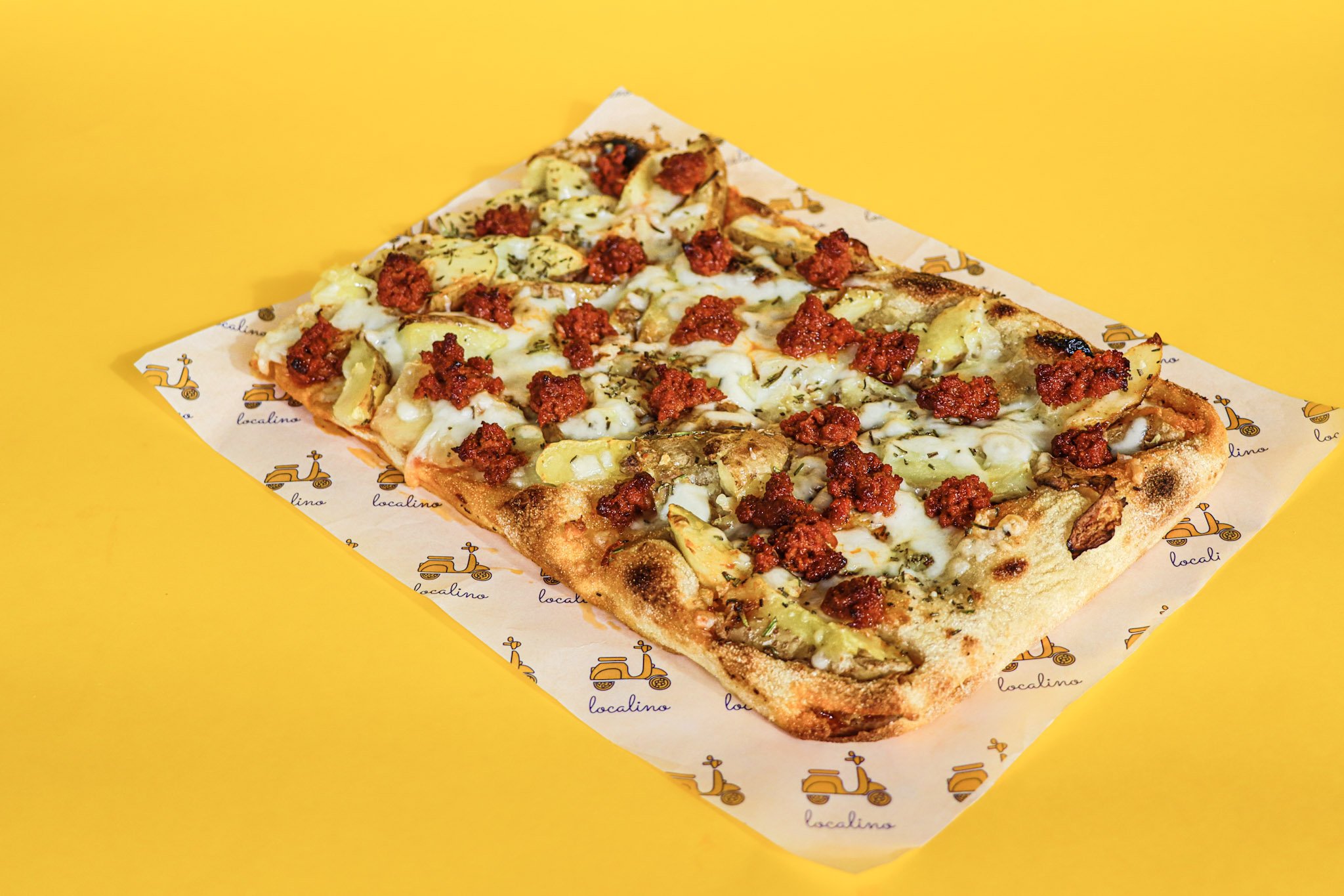 A rectangular slice of pizza topped with cheese, slices of potato, sausage crumbles, and herbs, placed on branded paper on a yellow background.