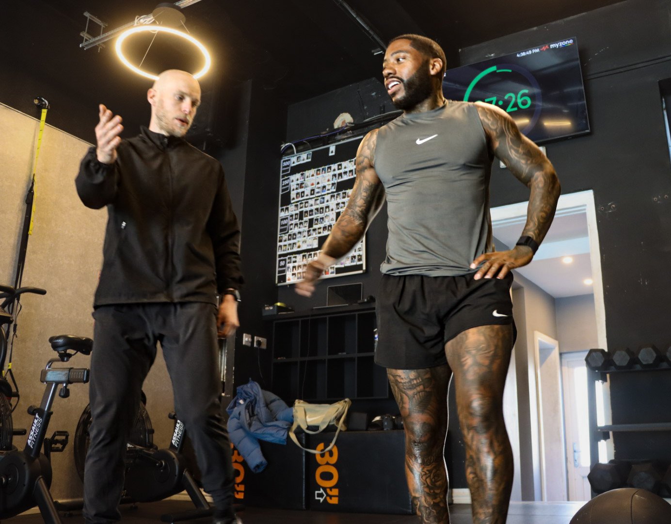 Two men in a gym, one wearing a black tracksuit and the other wearing a gray Nike sleeveless shirt and black shorts, engaging in conversation.